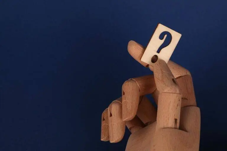 Wooden mannequin hand holding a block with a question mark against a blue background.