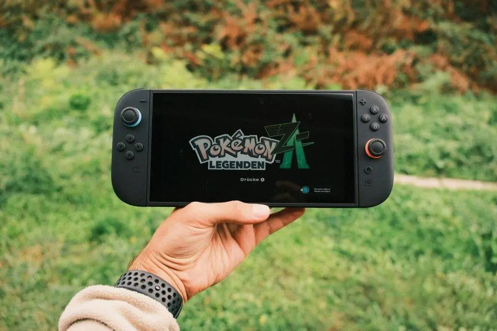 A hand holding a handheld gaming console displaying the Pok&eacute;mon Legends game screen outdoors.